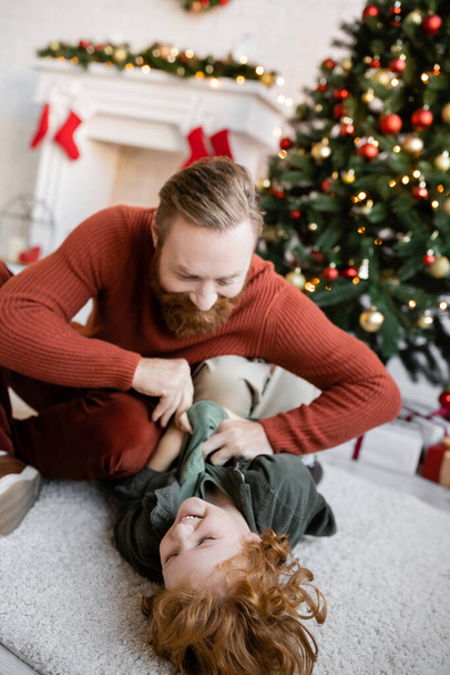redhead boy laughing on floor while having fun with dad near blurred Christmas tree - Foto, Imagem