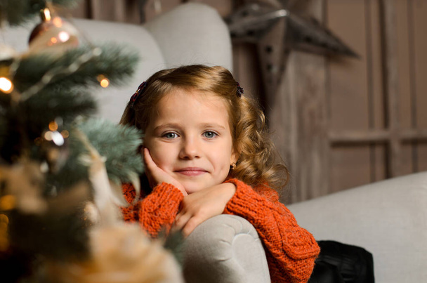 ένα όμορφο χαμογελαστό κορίτσι με ένα κόκκινο πουλόβερ κάθεται σε μια πολυθρόνα πίσω από ένα χριστουγεννιάτικο δέντρο, στηρίζοντας το πρόσωπό της με το χέρι της, με φόντο ένα ξύλινο τοίχο σοφίτα - Φωτογραφία, εικόνα