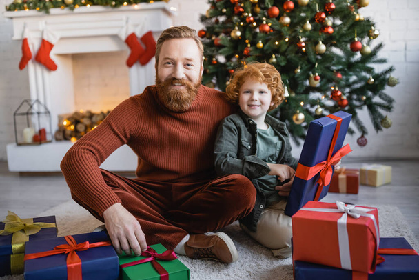 szakállas férfi vörös hajú gyerek mosolyog a kamera, miközben ül a padlón közelében ajándékdobozok és díszített karácsonyfa - Fotó, kép