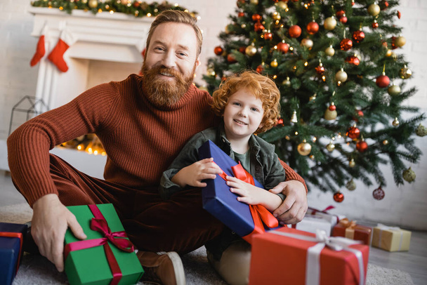 niño pelirrojo sonriente con papá barbudo mirando la cámara cerca de cajas de regalo y el árbol de Navidad en el fondo - Foto, imagen