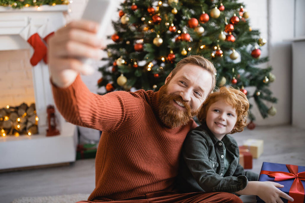 cheerful bearded man taking selfie with redhead son holding gift box near Christmas tree - Foto, afbeelding