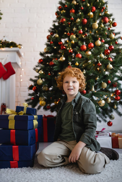 redhead boy sitting on floor near Christmas presents and pine tree decorated with baubles at home - Foto, Bild