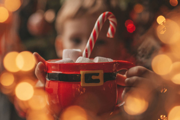 Happy kleine jongen dromer wachten op wonder Kerstman genieten van zoetigheid kleur lolly. Rood geruite slaapkledij vrolijk joch viert nieuwjaarsfeest kerstbeker met marshmallows bij noel tree eve 25 december - Foto, afbeelding