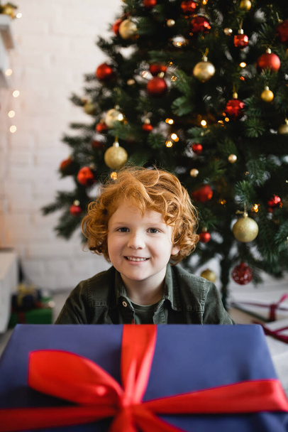 heureux garçon avec les cheveux rouges regardant la caméra près de la grande boîte cadeau et arbre de Noël sur fond flou - Photo, image