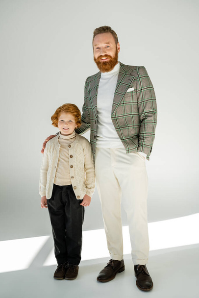 повна довжина модного бородатого чоловіка, що стоїть з рукою в кишені біля рудого сина на сірому фоні
 - Фото, зображення