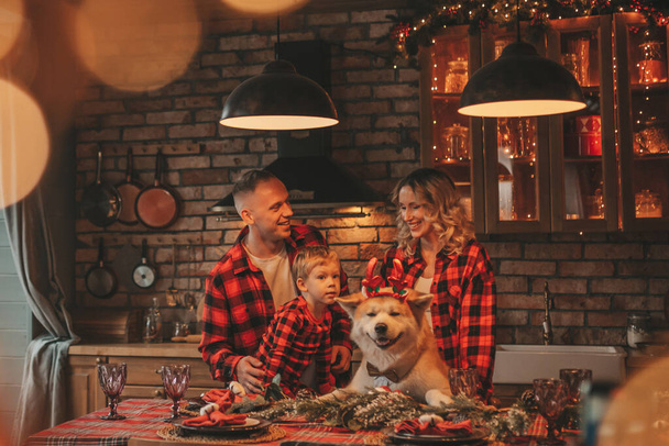 Πορτρέτο της ευτυχισμένης οικογένειας με το γιο σε κόκκινο καρό πιτζάμες γέλιο αγκαλιές φιλιά στο σπίτι παραμονή 25 Δεκέμβριος. Γιορτάζοντας το νέο έτος Noel ζεστό αγκαλιά ζεστού καιρού γιρλάντες φώτα αγκαλιάζει χαριτωμένο σκύλο του akita inu - Φωτογραφία, εικόνα