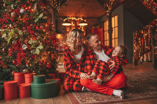 Χαμογελάστε δραστήριοι γονείς με μικρό γιο σε κόκκινο καρό υπνόσακους περιμένει Σάντα εσωτερική. Κομψή οικογένεια αγαπά γέλιο αγκαλιές φιλιά στο σπίτι. Γιορτάστε Πρωτοχρονιά γιρλάντες και φώτα παραμονή 25 Δεκέμβριος Noel δέντρο - Φωτογραφία, εικόνα