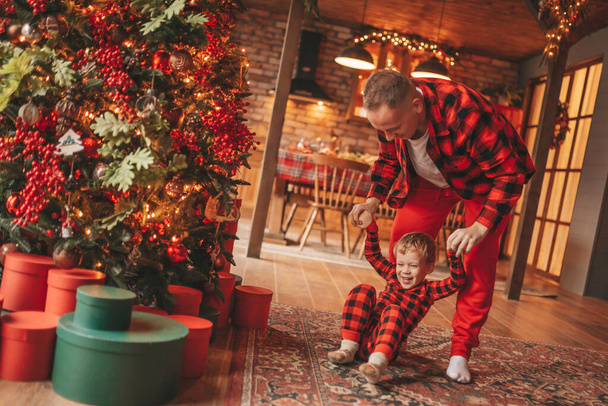笑顔の小さな男の子がお互いの屋内待っている奇跡と楽しんで父親の首に座っています。ノエルの木の冬の季節のガーランドライトで新年を祝うクリスマスの精神前夜25 12月父方の愛 - 写真・画像