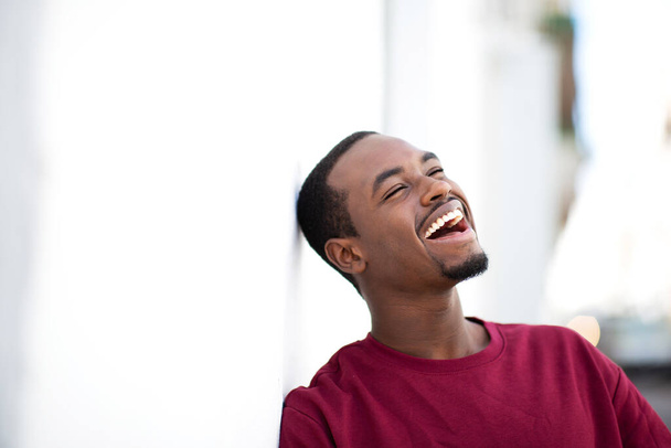 Closeup portrait of handsome young african man leaning to white wall smiling outside - Foto, imagen