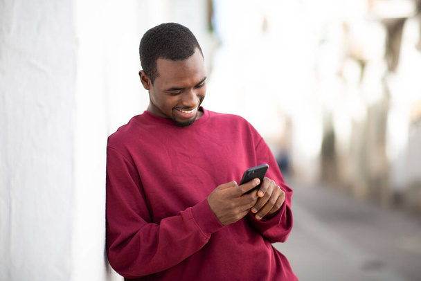 Портрет улыбающегося молодого чернокожего мужчины, прислонившегося к стене и переписывающегося на мобильном телефоне снаружи - Фото, изображение