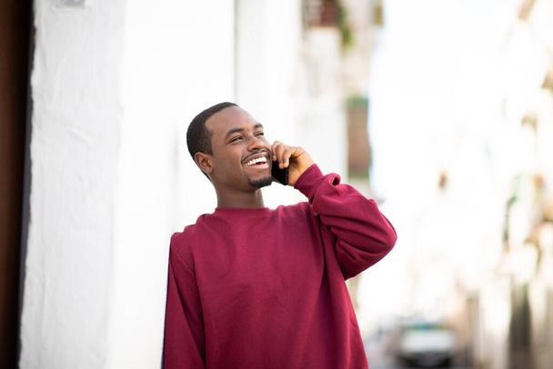 Portrait of happy young african american man leaning to wall and talking on smartphone outdoors in the city - Photo, Image