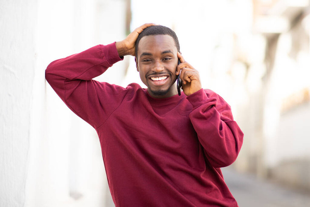 Porträt eines lächelnden schwarzen Mannes, der sich an die Wand lehnt und im Freien mit dem Handy spricht - Foto, Bild