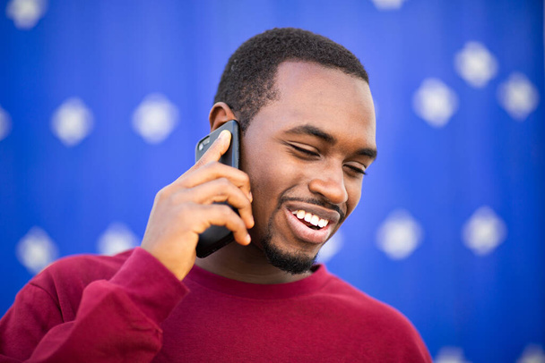 Nahaufnahme Porträt eines glücklichen Afrikaners, der auf seinem Handy vor blauem Hintergrund spricht - Foto, Bild