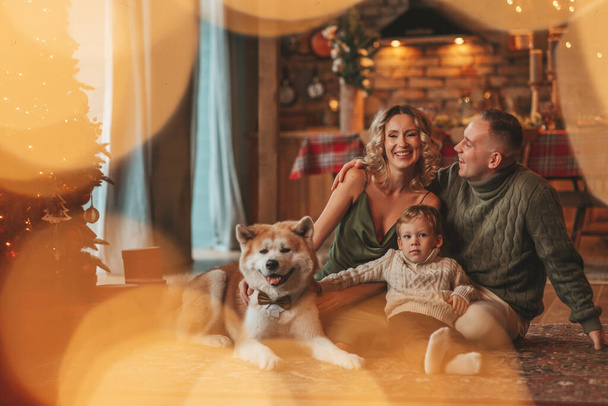 Porträt einer glücklichen Familie in gestrickten beigen Pullovern, die mit Akita Inu drinnen auf den Weihnachtsmann wartet. Lächelnde Eltern umarmen und küssen seinen kleinen Sohn Vorabend 25. Dezember feiert Weihnachten mit Lichtergirlanden Noelbaum - Foto, Bild