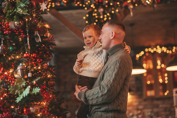 笑顔の男の子とお父さんはお互いの抱擁で楽しみ、屋内待っているサンタにキスをします。ノエルの木の冬の季節のガーランドライトで新年を祝うクリスマスの精神前夜25 12月父方の愛 - 写真・画像