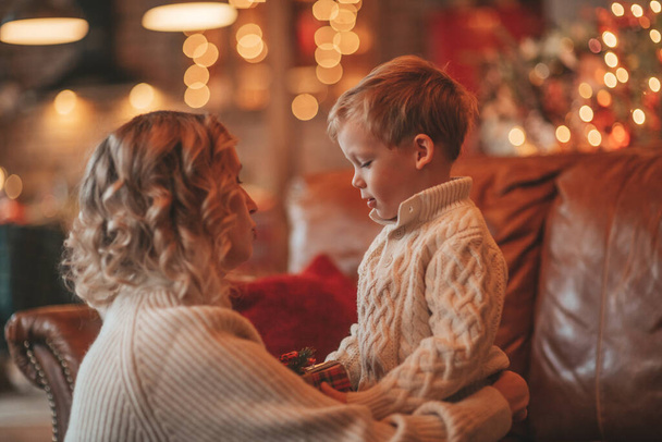 A boldog családi anya portréja kisfiúval kötött bézs pulóverben, ami a Mikulásra vár. Az anyák szeretik az öleléseket és a csókokat december 25-én ünneplik az újévi ajándékokat, melyek virágfüzért adnak Noel fának. - Fotó, kép