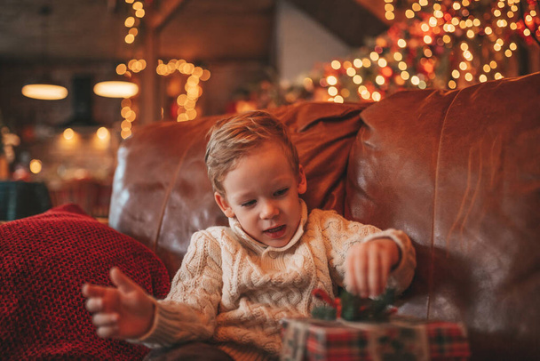 Smile small child being active having fun waiting for miracle Santa open presents. Cheerful kid in casual knit outfit celebrating new year with gifts boxes garlands bokeh lights noel eve 25 december - Фото, изображение