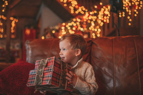 Hymyile pieni lapsi on aktiivinen hauskaa odottaa ihme Santa auki lahjoja. Iloinen lapsi rento neuloa asu juhlii uutta vuotta lahjat laatikot seppeleitä bokeh valot noel eve 25 joulukuu - Valokuva, kuva