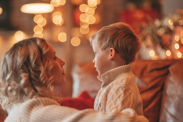 Muotokuva onnellinen perhe äiti pieni poika neulottu beige puserot odottaa Santa sisätiloissa. Äidit rakastavat halauksia ja suukkoja aattona 25 Joulukuu juhlia Uusi vuosi lahjoja antaa seppeleitä valot Noel puu - Valokuva, kuva