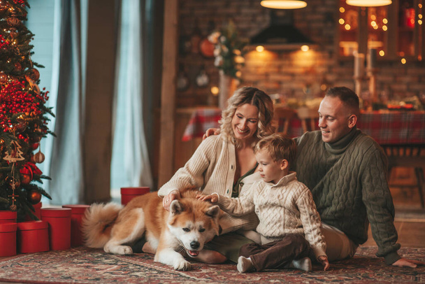 秋田犬とサンタ屋内を待っているニットベージュのセーターで幸せな家族の肖像画。笑顔の両親抱擁とキス彼の小さな息子eve 25 12月祝xmasとともにガーランドライト｜Noel tree - 写真・画像