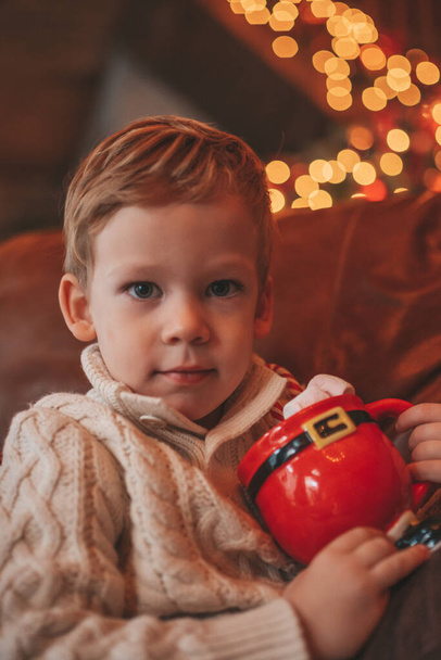 Happy little boy dreamer waiting at miracle Santa enjoy sweets color lollipop. Cheerful kid in knitwear outfit celebrating new year hold Xmas cup with marshmallows at bokeh lights noel eve 25 december - Photo, Image
