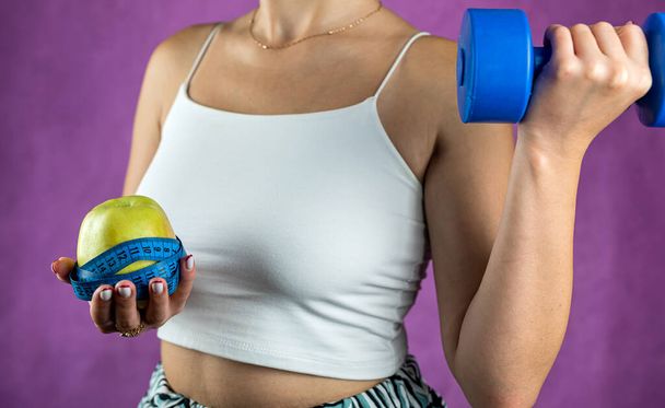 Slim unrecognizable woman with tape measure holding blue dumbbells and apple. Concept of weight loss and healthy lifestyle. - Photo, image