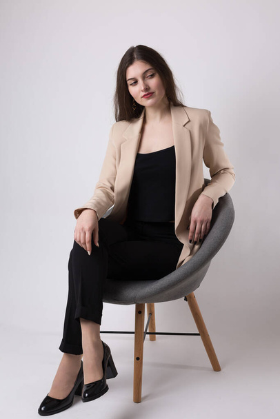 Portrait d'une jeune brune aux cheveux longs en studio. Jolie fille assise sur une chaise sur un fond blanc. - Photo, image