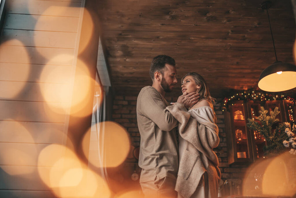 Portret młodej uroczej pary przytulającej się w kryty wieczór 25 grudnia. Kochankowie śmiejący się przytula pocałunki czekając na Boże Narodzenie w domu. Świętowanie nowego roku girlandy światła noel w eleganckim stroju dzianiny tkliwość - Zdjęcie, obraz