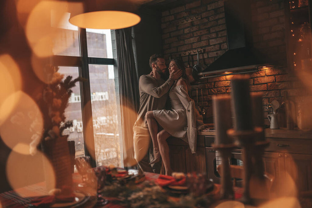 Portrét mladého krásného páru objímajícího se 25. prosince. Milenci se smějí, objímají a čekají doma na Vánoce. Oslava Nového roku věnce světla Noel v elegantním pletené oblečení jemnost - Fotografie, Obrázek