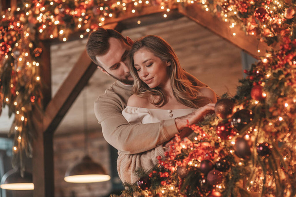 Portrait of young lovely couple hugging indoor eve 25 December. Lovers laughing hugs kisses waiting xmas at home. Celebrating new year garlands lights noel in elegant knitwear outfit tenderness - Foto, immagini