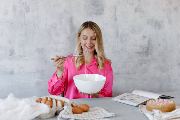 Femme souriante fouettant des œufs avec un fouet dans un bol de mélange blanc sur un fond gris. Une jeune femme blonde en chemisier rose prépare des pâtisseries dans la cuisine selon la recette du livre. - Photo, image