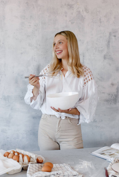 A happy woman whisking cream with a whisk in a white mixing bowl on a grey background.  Copy space.  - Фото, зображення