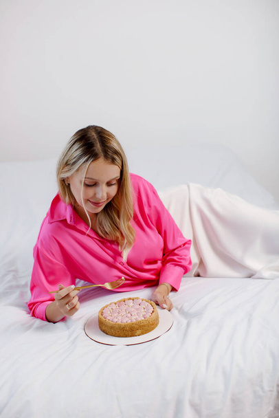 Beautiful young blonde woman in a pink blouse and white skirt eats a cheesecake with pink hearts in bed. Copy space. - Фото, зображення