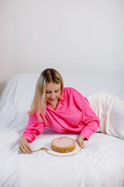 Kaunis nuori blondi nainen vaaleanpunainen pusero ja valkoinen hame syö juustokakku vaaleanpunainen sydämet sängyssä. Kopioi tila. - Valokuva, kuva