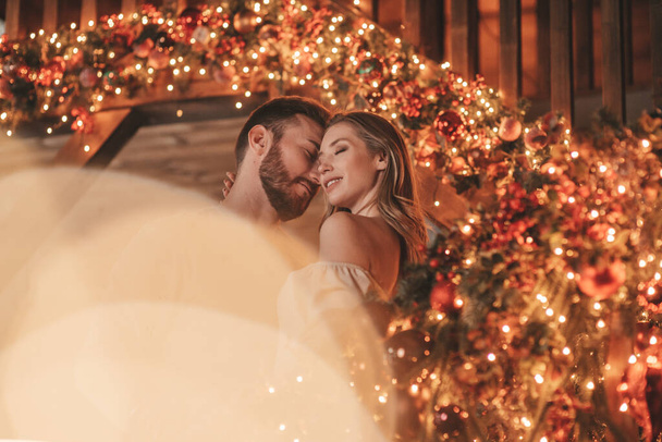 Portrait of young lovely couple hugging indoor eve 25 December. Lovers laughing hugs kisses waiting xmas at home. Celebrating new year garlands lights noel in elegant knitwear outfit tenderness - Фото, изображение