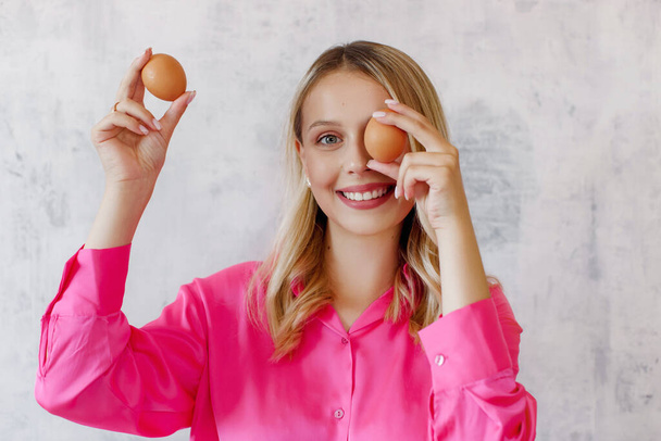 ピンクのブラウスの幸せな女性は灰色の背景のクローズアップで彼女の手の中に卵を保持しています。ブロンドの女性は台所で食べ物を準備します. - 写真・画像