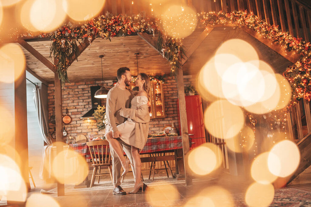 Muotokuva nuoresta ihanasta pariskunnasta halaamassa sisätiloissa 25. joulukuuta. Rakastavaiset nauravat halauksia odottamassa joulua kotona. Juhlitaan uusi vuosi seppeleitä valot noel tyylikäs neule asu arkuus - Valokuva, kuva