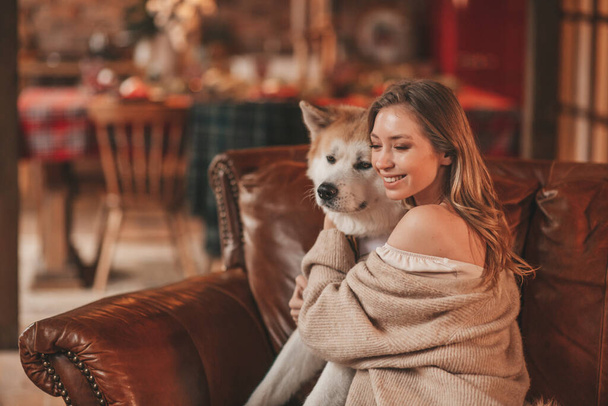 Portrait of young lovely couple hugging indoor eve 25 December with akita inu dog. People in knit stylish outfit hugs kisses tenderness celebrating new year garlands xmas lights noel tree at home - Valokuva, kuva