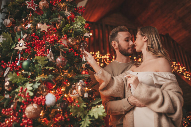 Portrait of young lovely couple hugging indoor eve 25 December. Lovers laughing hugs kisses waiting xmas at home. Celebrating new year garlands lights noel in elegant knitwear outfit tenderness - Φωτογραφία, εικόνα