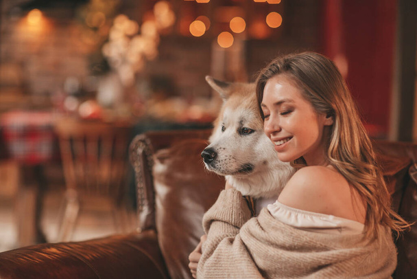 25 Aralık arifesinde evde Akia Inu köpeğiyle kucaklaşan genç bir çiftin portresi. Örgü ören şık elbiseli insanlar, yeni yılı kutlayan şefkat dolu öpücükler, Noel ışıkları, Noel ağacı. - Fotoğraf, Görsel