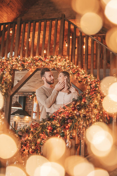 Portret młodej uroczej pary przytulającej się w kryty wieczór 25 grudnia. Kochankowie śmiejący się przytula pocałunki czekając na Boże Narodzenie w domu. Świętowanie nowego roku girlandy światła noel w eleganckim stroju dzianiny tkliwość - Zdjęcie, obraz