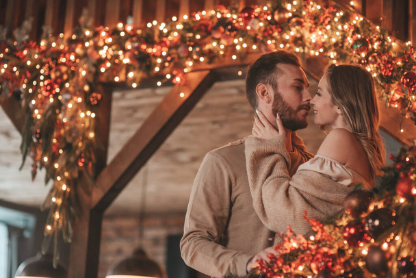 Portrait of young lovely couple hugging indoor eve 25 December. Lovers laughing hugs kisses waiting xmas at home. Celebrating new year garlands lights noel in elegant knitwear outfit tenderness - Foto, afbeelding