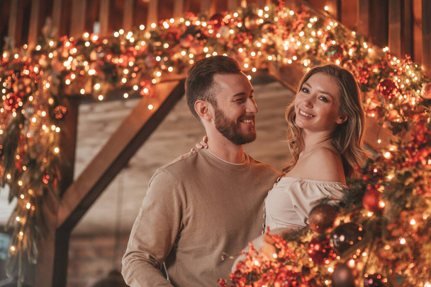Muotokuva nuoresta ihanasta pariskunnasta halaamassa sisätiloissa 25. joulukuuta. Rakastavaiset nauravat halauksia odottamassa joulua kotona. Juhlitaan uusi vuosi seppeleitä valot noel tyylikäs neule asu arkuus - Valokuva, kuva