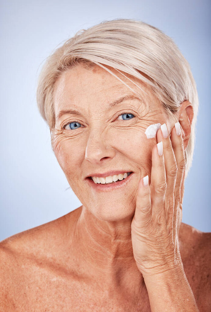 Portrait, skincare and senior woman with face cream for moisturizing on blue studio background. Beauty, smile or elderly female model apply facial creme, cosmetics product or lotion for healthy skin. - Foto, Imagen