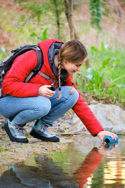 Νεαρό θηλυκό πεζοπόρος γεμίζει το νερό μπουκάλι του ακατέργαστου νερού σε ένα ρυάκι. - Φωτογραφία, εικόνα