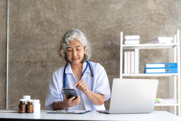 Доктор старший седовласый женщина в белом медицинском халате использует смартфон и улыбается во время сидеть использовать ноутбук в офисе. - Фото, изображение