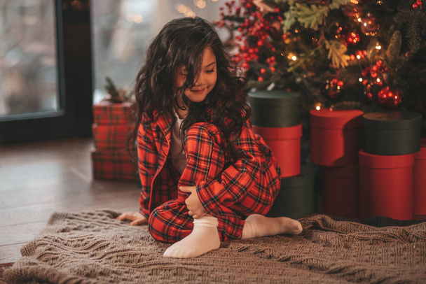 Szczęśliwy japoński mała dziewczynka marzycielka czeka cud od Mikołaja pozowanie na podłodze w pobliżu noel drzewa. Azjatycki dzieciak w czerwonej kratę strój świętuje nowy rok z pudełek na prezenty bokeh xmas światła przeddzień 25 Grudzień - Zdjęcie, obraz