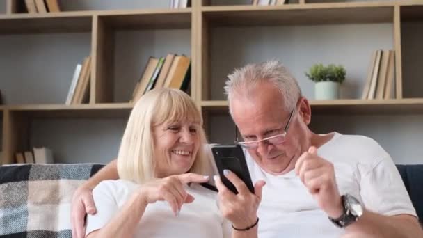 Szczęśliwi emerytowani dziadkowie relaksujący się w domu na sofie i przy użyciu nowoczesnego smartfona. Osoby starsze korzystają z Internetu - Materiał filmowy, wideo