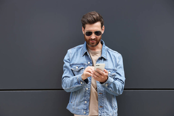 Handsome bearded man with smartphone near grey wall outdoors - 写真・画像