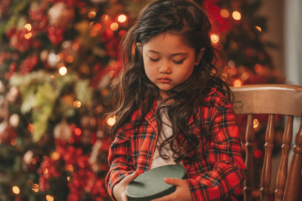 Heureuse petite fille japonaise rêveuse attendant miracle du Père Noël posant sur le sol près de noel tree. Enfant asiatique en costume à carreaux rouges célébrant la nouvelle année avec des boîtes-cadeaux à bokeh lumières de Noël veille 25 décembre - Photo, image